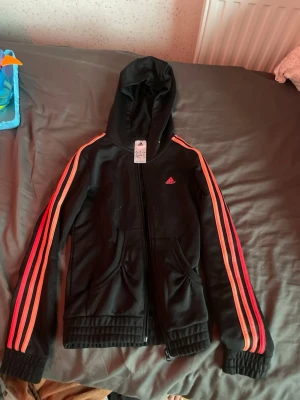 Svart Adidas hoodie med rosa ränder - Svart hoodie från Adidas med dragkedja och huva. Klassiska rosa och orangea ränder längs ärmarna och Adidas-logga på bröstet. Mjukt material och två fickor framtill. Perfekt för en sportig och avslappnad stil. 11-12 år 