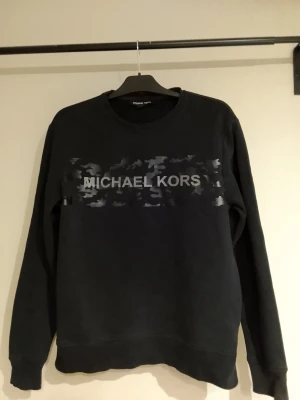 Svart sweatshirt Michael Kors NYSKICK, FYND! - Svart sweatshirt från Michael Kors med rund halsringning och tryckt logga framtill i grått över ett camo-inspirerat mönster. Långärmad och i klassisk, avslappnad passform. Perfekt för en stilren och trendig look. Nyskick, riktigt FYND!! ÄKTA Michael Kors 👌🏼🤌🏼