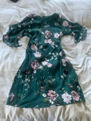 Grön blommig klänning med volangärm - Grön klänning med rosa och vita blommor, trekvartsärm med volang och rund halsringning. Mjuk och lätt i materialet, perfekt för varma dagar. Snygg passform som ger en feminin siluett.