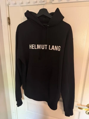 Svart Helmut Lang hoodie med tryck - Svart hoodie från Helmut Lang med vit logotyp på bröstet. Klassisk modell med huva och känguruficka. Tillverkad i 100% bomull med detaljer i bomull och elastan för extra komfort. Perfekt för en stilren och avslappnad look.