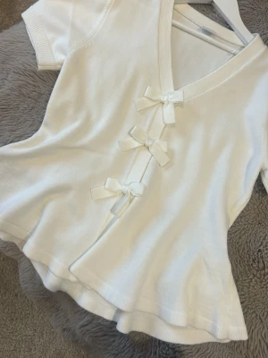 Vit omlottopp med rosetter - Supersöt vit omlottopp med tre dekorativa rosetter framtill och v-ringning. Toppen har korta ärmar och är tillverkad i ett mjukt, stickat material som ger en feminin och trendig look.