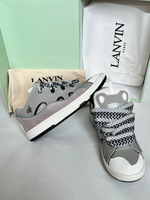 Grå Lanvin sneakers med mönstrade snören - Lanvin sneakers i grått och vitt med coola, breda snören i svartvitt zigzag-mönster. Skorna har en chunky sula, detaljer i mocka och läder samt vit tåhätta. Märkeslogga på sidan och plös. Perfekta för dig som gillar statement-sneakers.