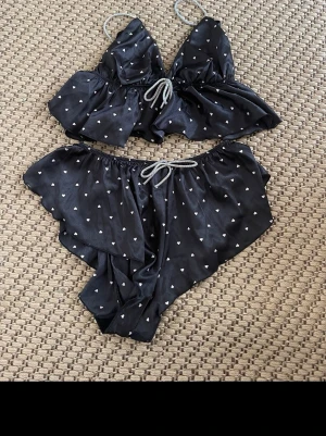 Svart pyjamasset med hjärtmönster - Sött tvådelat pyjamasset i svart satin med små vita hjärtan. Setet består av ett linne med smala axelband och volangkant samt matchande shorts med resår och knytband i midjan. Perfekt för mysiga kvällar hemma. (Har en sänerhetsnåll så den sitter bättre)