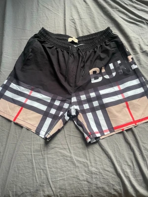 Svarta och rutiga Burberry shorts - Säljer ett par snygga shorts/badshorts från Burberry med klassiskt rutigt mönster i svart, beige, vitt och rött. Shortsen har snörning i midjan, sidofickor och en bakficka med knapp. Perfekta för stranden eller poolen. 