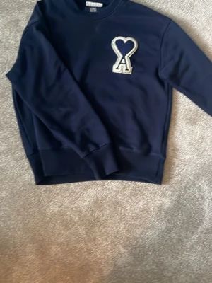 Mörkblå sweatshirt från Ami Paris - Mörkblå sweatshirt från Ami Paris med deras ikoniska hjärta-A-logga i vitt och blått på bröstet. Tröjan har rund halsringning, ribbade muddar och är gjord i mjukt bomullsmaterial. Perfekt för en avslappnad och trendig stil.