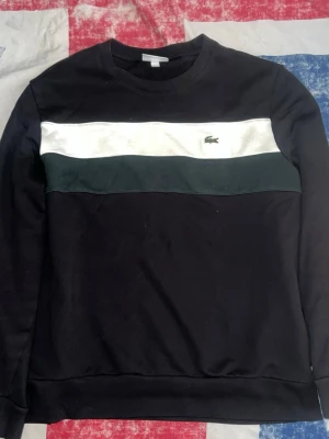 Svart sweatshirt från Lacoste - Cool svart sweatshirt från Lacoste med vita och mörkgröna paneler över bröstet. Klassisk rund halsringning och broderad krokodil-logga på bröstet. Perfekt för en avslappnad och stilren look.