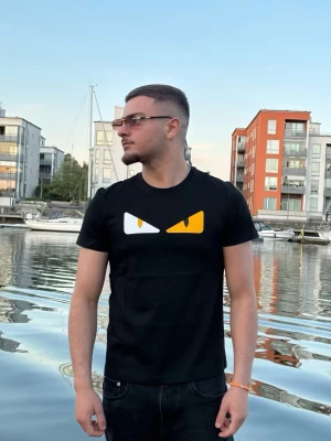 Svart Fendi t-shirt med ögontryck - Svart Fendi t-shirt med ett coolt tryck av två ögon i vitt och gult på bröstet. Klassisk passform och rund halsringning. Perfekt för dig som vill sticka ut lite extra med din stil.
