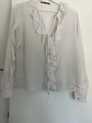 Vit volangblus från Zara - Snygg vit blus från Zara med volangdetaljer längs knäppningen och kragen. Blusen har långa ärmar med manschett och är tillverkad i ett tunt, lätt och transparent tyg som ger en luftig känsla. Perfekt för att skapa en romantisk och trendig look.
