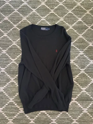 Svart stickad tröja Polo Ralph Lauren - Klassisk svart stickad tröja från Polo Ralph Lauren med rund halsringning och den ikoniska röda logotypen broderad på bröstet. Tröjan har långa ärmar och ribbade muddar vid ärmslut och nederkant. Perfekt till lager på lager och enkel att matcha.