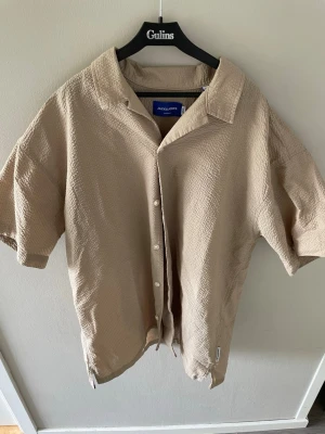 Beige kortärmad skjorta Jack & Jones - Snygg beige skjorta från Jack & Jones med korta ärmar och avslappnad passform. Skjortan har en krage, knäppning framtill och är tillverkad i ett strukturerat bomullstyg. Perfekt för en chill och stilren look.