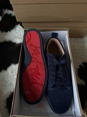 Mörkblå Louboutin sneakers - Snygga mörkblå sneakers från Christian Louboutin med klassisk röd sula och diskret logga på hälen. Skorna är i mocka med blåa snören och blåa detaljer. Perfekta för dig som vill sticka ut med en lyxig och stilren look fina.