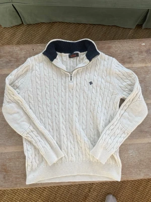 Vit Morris kabelstickad tröja med halvzip - Säljer en vit kabelstickad tröja från Morris med halvzip och mörkblå krage. Tröjan har ett broderat emblem på bröstet och ribbstickade muddar. Perfekt för dig som gillar klassisk och stilren look.