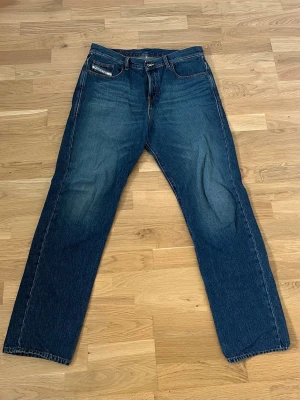 Blå raka jeans med klassisk passform från Diesel - Säljer ett par blå jeans med rak passform och klassisk femficksdesign. Jeansen har diskreta slitningar framtill och kontrastsömmar. Perfekta för en avslappnad stil. Materialet är denim i bomull. Byxorna är från Diesel