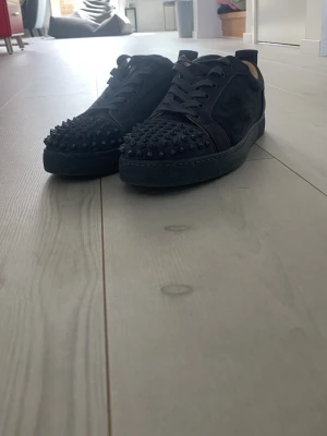 Svarta Louboutin sneakers med nitar - Säljer ett par svarta sneakers från Louboutin med coola svarta nitar på tån och sidorna. Skorna har en klassisk låg siluett, snörning och är tillverkade i mocka och läder. Perfekta för dig som vill sticka ut med en lyxig och edgy stil.