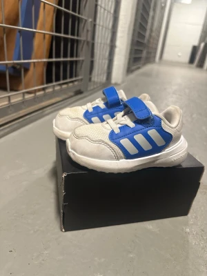 Barn skor sneakers från Adidas - Fräscha sneakers från Adidas i vitt och blått med klassiska tre ränder på sidan. Skorna har mesh- och syntetmaterial, kardborreband och elastiska snören för enkel påtagning. Perfekta för en sportig och avslappnad stil.