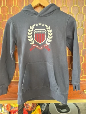 Mörkblå hoodie från GANT med tryck - Snygg mörkblå hoodie från GANT med stort tryck framtill i vitt och rött, prydd med logga, stjärnor och lagerkrans. Klassisk känguruficka och ribbade muddar. Perfekt för en avslappnad och sportig stil.