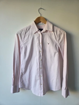 Rosa randig skjorta från Gant - Klassisk rosa och vit randig skjorta från Gant med lång ärm och knappar framtill. Skjortan har en liten broderad logga på bröstet och är tillverkad i bomull. Perfekt för en clean och stilren look.