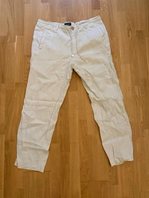 Beiga linnebyxor från White Linen Athens - Snygga beiga linnebyxor från White Linen Athens med dragsko och knapp i midjan. Byxorna har raka ben, sidfickor och är tillverkade i ett luftigt linnematerial som passar perfekt för en avslappnad stil.