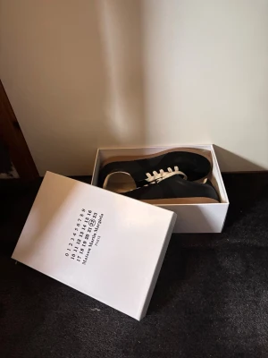 Svarta sneakers Maison Margiela - Stilrena svarta sneakers från Maison Margiela med vita snören och beige sula. Skorna har en klassisk låg siluett och är tillverkade i en mix av läder och mocka. Perfekta för dig som gillar minimalistisk och trendig design.