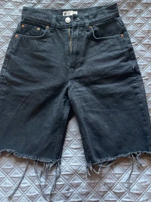 Svarta jeansshorts med rå kant - Svarta jeansshorts från Gina Tricot med hög midja och rå, fransig kant nertill. Klassisk femficksmodell med dragkedja och knapp framtill. Perfekta för en avslappnad och trendig look.
