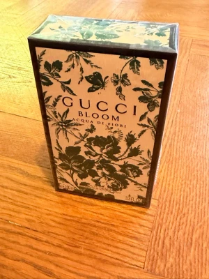 Gucci Bloom Acqua di Fiori parfym - Gucci Bloom Acqua di Fiori parfym i en elegant rektangulär kartong med grön botanisk design på beige bakgrund. Förpackningen har tydlig logotyp och stilrena detaljer som ger en lyxig känsla. Volymen är 100 ml.