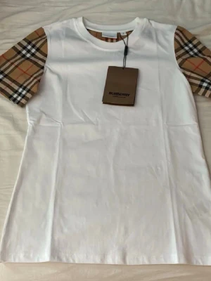 Vit Burberry t-shirt med rutiga ärmar - Snygg vit t-shirt från Burberry med klassiska rutiga ärmar i beige, svart och rött. T-shirten har rund halsringning och är tillverkad i mjuk bomull. Perfekt för dig som vill ha en stilren look med en touch av lyx.