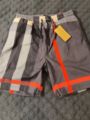 Grå-rutiga shorts från Burberry - Säljer ett par snygga shorts från Burberry med klassiskt rutigt mönster i grått, svart, vitt och röda detaljer. Shortsen har snörning i midjan och är tillverkade i ett lätt och bekvämt material, perfekta för sommaren. PASSAR M-L. HELT NY, OANVÄND.