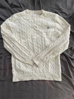 Vit kabelstickad tröja från Gant - Kabelstickad vit tröja från Gant med rund halsringning och diskret logga broderad på bröstet. Tröjan har långa ärmar och ribbade muddar vid ärmslut och nederkant. Perfekt för en stilren och klassisk look.