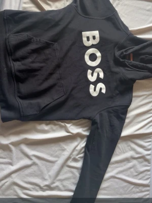 Svart hoodie från BOSS med tryck - Svart hoodie från BOSS med stor vit logga på bröstet. Klassisk känguruficka framtill och huva med dragsko. Mjuk bomullskänsla och avslappnad passform, perfekt för en chill och stilren look.