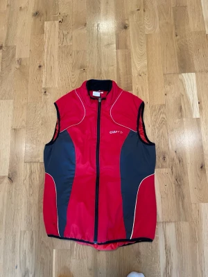 Röd sportväst från Craft stl 38 fint skick - Sportig röd väst från Craft med svarta paneler på sidorna och vita detaljer. Västen har dragkedja framtill och står ut med sin atletiska design, perfekt för aktiva dagar. Materialet känns lätt och funktionellt.