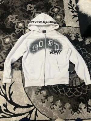 Vit hoodie med tryck och dragkedja - Snygg vit hoodie med svart och vitt tryck på bröstet där det står 'Hood Rich'. Dragkedja framtill, två fickor och huva med textdetalj. Perfekt för en streetwear-look. Mjuk insida och ribbade muddar. Passar dig som gillar en avslappnad stil.