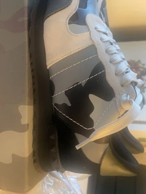 Valentino rock runner - Snygga sneakers med grått, svart och vitt kamouflagemönster. Skorna har vita snören, svarta sulor och detaljer i mocka och läderimitation. Perfekt för dig som vill sticka ut med en cool streetstyle-look.
