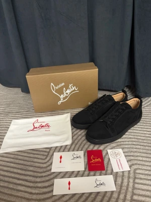 Svarta Christian Louboutin sneakers - Snygga svarta sneakers från Christian Louboutin med klassisk röd sula och diskret logga på sidan. Skorna har lågt skaft, svarta skosnören och är tillverkade i en mix av läder och mocka. Perfekta för dig som vill sticka ut med exklusiv stil.