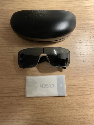 Versace Y2K Vintage Solglasögon MOD 2037 - Versace Mod 2037 1001/87 solglasögon med svart metallbåge och gra linser i wraparound-modell. Mindre, ytliga repor - knappt anvanda - samt något lösa skalmar som enkelt kan justeras och dras åt igen. I övrigt i mycket gott skick. Levereras med originalfodral.