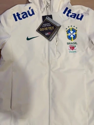 Vit Brasil Nike vindjacka GORE-TEX - STORLEK M MEN PASSAR FÖR DE SOM HAR S (LITEN I STORLEK) Vit vindjacka från Nike med Brasil CBF-logga, blåa Itau-tryck på axlarna och Guaraná-logga. Jackan har huva, hel dragkedja och är tillverkad i GORE-TEX-material som står emot regn och vind. Perfekt för dig som gillar sportig stil och vill sticka ut.