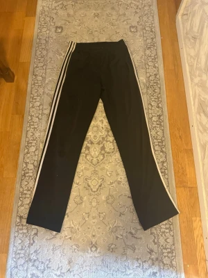 Svarta Adidas mjukisbyxor med ränder - Svarta mjukisbyxor från Adidas med klassiska vita tre ränder längs båda benen. Byxorna har rak passform och resår i midjan. Perfekta för en avslappnad och sportig stil. Materialet är mjukt och stretchigt för extra komfort. Fläckar vid nederkant av benen