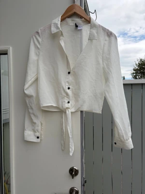 Vit knytblus med krage H&M - Stilren vit blus från H&M med klassisk krage, knappar framtill och knytning i midjan. Långärmad modell med svarta knappar vid ärmslut och framtill. Perfekt att matcha med jeans eller kjol för en trendig look. storlek 34 xs men kan även passa  36.