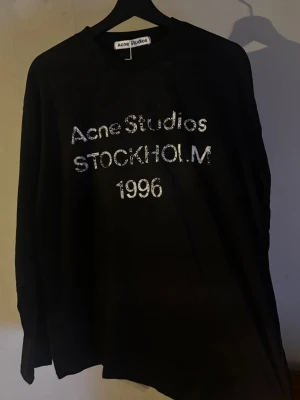 Svart långärmad t-shirt Acne Studios - Svart långärmad t-shirt från Acne Studios med tryck i vitt framtill där det står 'Acne Studios STOCKHOLM 1996'. Klassisk passform och rund halsringning. Perfekt för en stilren och trendig look.