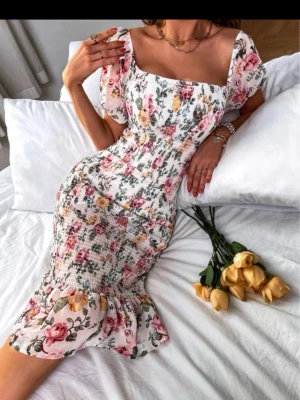 Blommig sommarklänning med volang - Supersöt blommig klänning med smockad midja, volangkant nertill och puffärmar. Klänningen har en vit bas med rosa, gula och gröna blommor och fyrkantig ringning. Perfekt för varma dagar och ger en romantisk vibe.