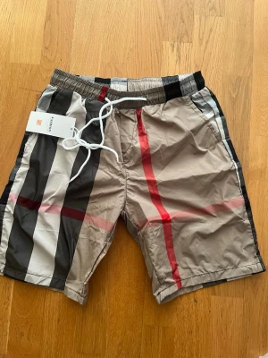 Birberry shorts - Snygga beige shorts från Burberry med klassiskt rutigt mönster i svart, vitt och rött. Shortsen har elastisk midja med vit snörning och två sidofickor. Perfekta för sommaren och ger en trendig look.