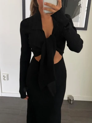 Svart långklänning med knyt och krage - ADOORE STRL M/38 - Svart långklänning med lång ärm, krage och snygg knytning framtill som skapar en cool cut-out detalj vid midjan. Klänningen har en figurnära passform och är ribbad för extra struktur. Perfekt för dig som gillar trendiga och stilrena plagg. Passar på mig som är i storlek 34/36