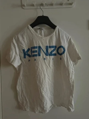 Vit kenzo tröja från Paris med blå logga - Vit t-shirt från Kenzo med stor blå logotyp på bröstet. Klassisk rund halsringning och korta ärmar. Tillverkad i mjuk bomull som känns skön mot huden. Perfekt för en avslappnad och trendig look.