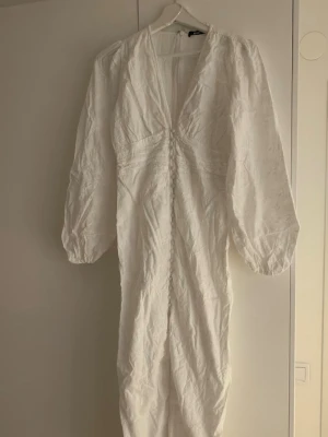Vit långklänning från Gina Tricot - Säljer en vit långklänning från Gina Tricot med v-ringning och långa puffärmar. Klänningen har dekorativa knappar framtill och ett lätt glansigt tyg med subtilt mönster. Perfekt för dig som gillar en romantisk och trendig stil. Klänningen är använd 1 gång och skulle säga att den passar om man har en mindre 36🌸🩷