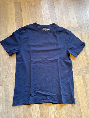 Mörkblå t-shirt från Polo Ralph Lauren - Klassisk mörkblå t-shirt från Polo Ralph Lauren med röd broderad logga på bröstet. T-shirten har rund halsringning och korta ärmar. Tillverkad i mjuk bomull för en skön känsla och enkel stil.