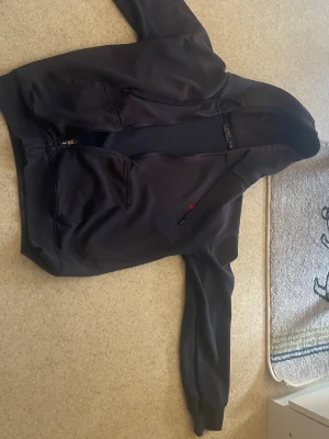 Zip hoodie från Polo Ralph Lauren - Klassisk svart hoodie från Polo Ralph Lauren med dragkedja, huva och två stora fickor framtill. Liten röd logga broderad på bröstet. Tillverkad i mjukt bomullsmaterial som är skönt att bära. Perfekt för en avslappnad och stilren look.