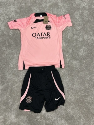 PSG rosa och svarta fotbollskläder - Säljer ett PSG fotbollskit med en rosa t-shirt och svarta shorts med rosa detaljer. Tröjan har PSG-logga, Nike-logga och Qatar Airways-tryck. Shortsen har elastisk midja och PSG-märke. Perfekt för fotbollsträning eller match.