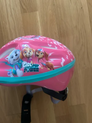 Rosa Paw Patrol cykelhjälm barn - Säljer en rosa cykelhjälm med motiv från Paw Patrol och texten 'Girl Pup Power'. Hjälmen har justerbara remmar och vit insida med svart vaddering. Perfekt för dig som vill ha en färgglad och lekfull hjälm till cykelturen.