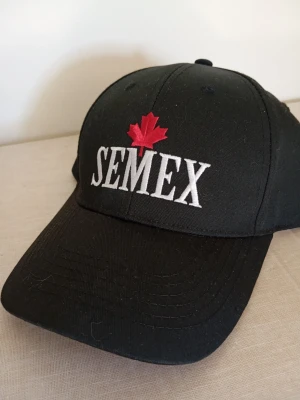 Svart keps med SEMEX-logga - Svart keps med böjd skärm och broderad vit SEMEX-text framtill samt ett rött lönnlöv ovanför loggan. Klassisk modell med justerbar baksida. Perfekt accessoar för en sportig och avslappnad stil.