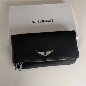Svart kuvertväska Zadig & Voltaire - Snygg svart kuvertväska i läder från Zadig & Voltaire med silverfärgad dragkedja och ikoniskt vingemblem framtill. Stilren och modern design med struktur i materialet. Perfekt accessoar för att lyfta din outfit.