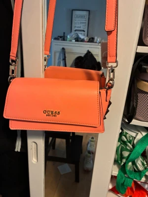 Korallrosa handväska från Guess - Snygg korallrosa handväska från Guess med guldfärgad logga på framsidan. Väskan har en rektangulär form, justerbar axelrem och detaljer i metall. Insidan är ljus med Guess-mönster och en liten metallplatta med logga. Perfekt för att lyfta din outfit.